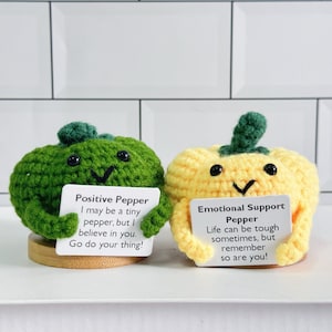 May include: Two crocheted pepper plushies, one green and one yellow, holding signs with positive messages. The green pepper says "Positive Pepper I may be a tiny pepper, but I believe in you. Go do your thing!" The yellow pepper says "Emotional Support Pepper Life can be tough sometimes, but remember so are you!"