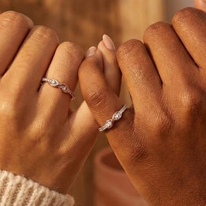 Peut inclure: Deux mains portant des bagues. L'une des bagues présente une pierre centrale ovale avec de petites pierres de chaque côté. L'autre bague a une série de petites pierres. Les mains portent un pull crème et un pull rouille.