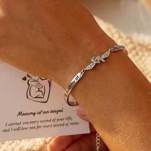 May include: A silver bracelet with the words "Mommy of" engraved on it. The bracelet features a charm with angel wings and small crystals. A card with the text "Mommy of an angel" is held in the background.
