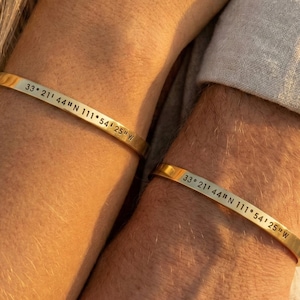 Può includere: Due braccialetti in metallo color oro, ciascuno inciso con le coordinate "33° 21' 44" N 111° 54' 25" W". I braccialetti sono semplici fasce, indossate ai polsi. L'immagine mostra un primo piano dei braccialetti.