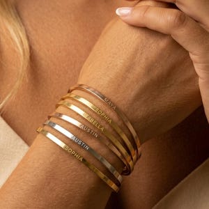 May include: A stack of gold and silver-toned bangles, each engraved with a name. The names include "Maya", "Sophia", "Isabella", and "Austin". The bracelets are worn on a wrist, showcasing the personalized jewelry.