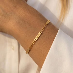 May include: Gold-colored bracelet with a rectangular nameplate that reads "Connor" in a vertical orientation. The bracelet is made of small, linked chain segments. The bracelet is worn on a wrist.