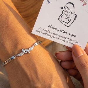May include: A silver bracelet with the words "Mommy of" engraved on it. The bracelet features a charm with baby footprints and angel wings. A card with the text "Mommy of an angel" and a mother and child illustration is held nearby.