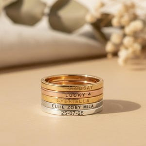 Peut inclure: Une pile de bagues personnalisées en or, or rose et argent. Chaque bague est gravée d'un nom ou d'une date. Les bagues sont empilées, créant un effet superposé. Le fond est de couleur neutre et douce.