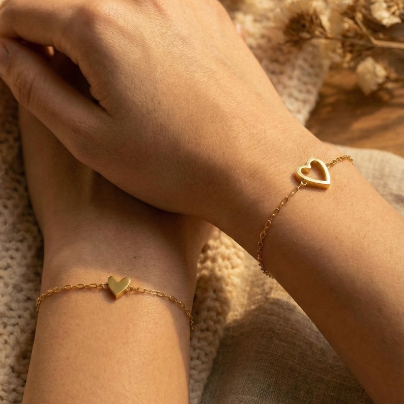 May include: Two gold-colored heart charm bracelets. One bracelet features a solid heart charm, while the other has an open heart design. The bracelets are on delicate chain links. The background is a soft, neutral color.
