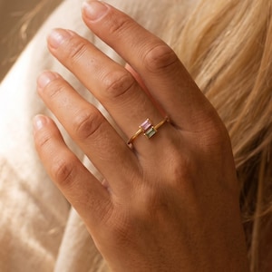 Könnte beinhalten: Ein goldfarbener offener Ring mit zwei im Baguette-Schliff geschliffenen Edelsteinen, einer in einem zarten Rosaton und der andere in einem gedämpften Grünton, auf einer dünnen, polierten Metallschiene, die sich um den Finger legt.
