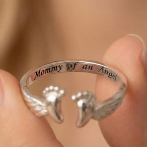 Könnte beinhalten: Ein silberner Ring mit der Aufschrift "Mommy of an Angel". Der Ring hat ein Design aus Engelsflügeln und Babyfüßen. Der Ring wird in der Hand einer Person gehalten.