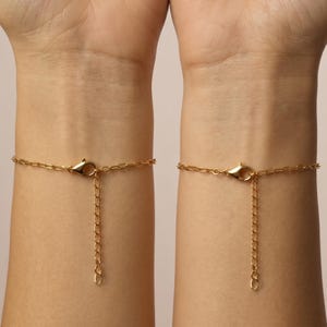 May include: Two gold-colored chain bracelets, each with a lobster clasp and an adjustable chain. The bracelets are shown on wrists against a neutral background. The chains have a delicate, linked design.