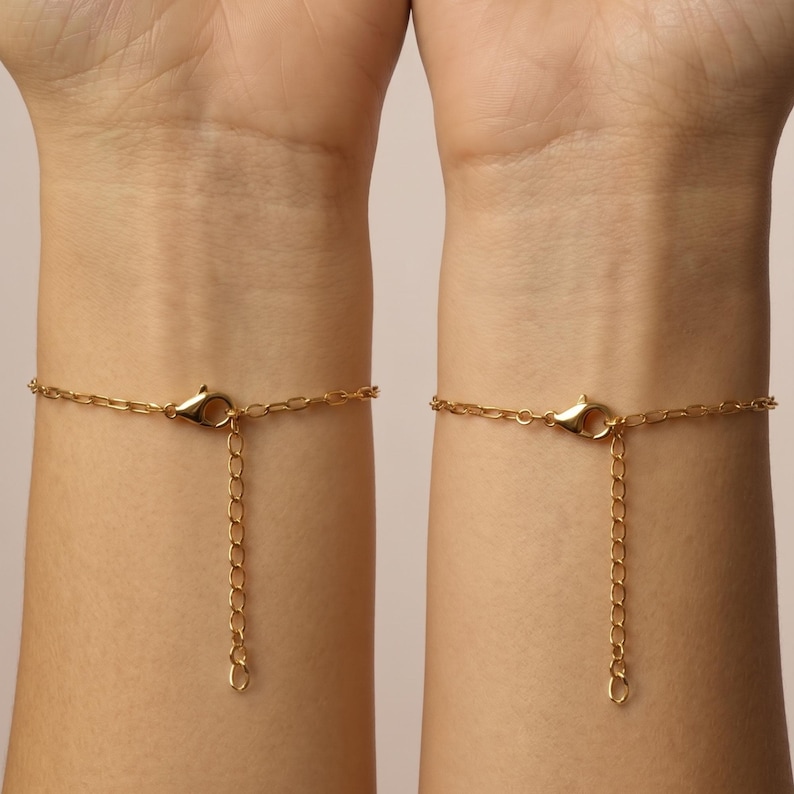 May include: Two gold-colored chain bracelets, each with a lobster clasp and an adjustable chain. The bracelets are shown on wrists against a neutral background. The chains have a delicate, linked design.