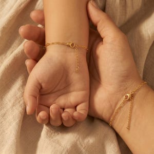 May include: Two gold-colored chain bracelets, one on a child's wrist and one on an adult's wrist. The bracelets have a delicate chain design with a lobster clasp closure and an adjustable chain. The background is a neutral beige fabric.