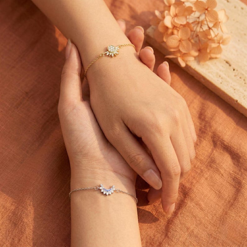 May include: Two delicate bracelets are displayed in this image. One is gold with a sunburst design, and the other is silver with a crescent moon design. The bracelets are on wrists, with a warm, neutral background.