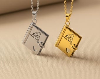May include: Two necklaces with book-shaped pendants. One pendant is silver, the other gold. Each pendant has a heart-shaped clasp and a design on the cover. The chains are delicate and the background is a neutral beige.