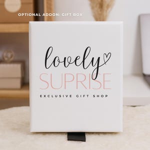 May include: A white gift box with the words "lovely surprise" in black and pink script. The text "exclusive gift shop" is below. A black ribbon is attached to the bottom of the box. The words "optional addon: gift box" are at the top.