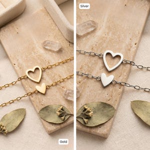 May include: Two delicate heart-shaped bracelets, one gold and one silver, are displayed. Each bracelet features a chain with a larger open heart and a smaller solid heart charm. The bracelets rest on a textured surface with dried leaves.