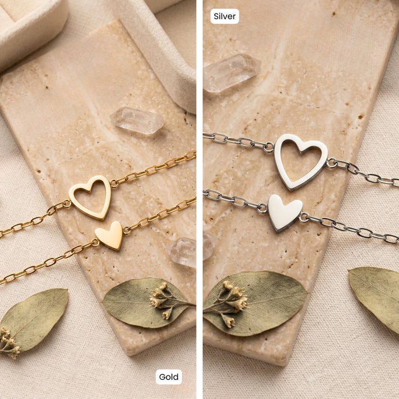May include: Two delicate heart-shaped bracelets, one gold and one silver, are displayed. Each bracelet features a chain with a larger open heart and a smaller solid heart charm. The bracelets rest on a textured surface with dried leaves.