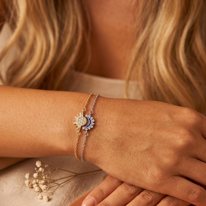 May include: A gold and silver bracelet featuring sun and moon charms. The sun charm is gold with clear and white stones, while the moon charm is silver with blue stones. The bracelet is worn on a wrist.