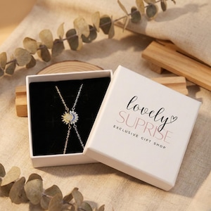 May include: A silver necklace with a sun and moon pendant, displayed in an open white gift box with a black interior. The box has the words "lovely SURPRISE" printed on the lid. The necklace is on a neutral-toned surface.