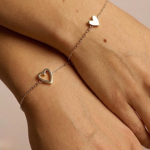 May include: Two silver bracelets, each featuring a heart charm. The bracelets are linked by a delicate chain and are displayed on wrists. The heart charms are the focal point, adding a touch of elegance to the jewelry.