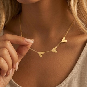 Peut inclure: Un collier doré avec trois pendentifs en forme de cœur. Chaque cœur porte un nom gravé : Chloe, Leo et Mia. Le collier est présenté, mettant en valeur la chaîne fine et le design en forme de cœur.