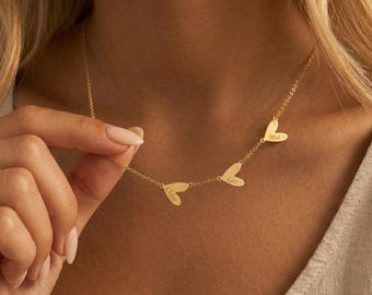 Könnte beinhalten: Eine goldfarbene Halskette mit drei herzförmigen Anhängern. Jeder Anhänger trägt einen eingravierten Namen: Chloe, Leo und Mia. Die Halskette wird präsentiert, wobei die feine Kette und das Herzdesign hervorgehoben werden.