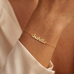 May include: A delicate gold bracelet with the name "Isabella" in cursive script. The bracelet is worn on a wrist, with a soft, white fabric in the background. The bracelet is a personalized jewelry item.