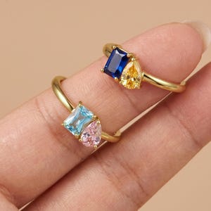 May include: Two gold rings, each featuring a dual-stone design. One ring displays a rectangular blue stone and a pear-shaped yellow stone. The second ring features a rectangular light blue stone and a pear-shaped pink stone.