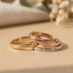 Puede incluir: Tres anillos personalizados en oro, oro rosa y plata. El anillo dorado dice "LUCKY" con un símbolo de trébol. El anillo de oro rosa dice "LINDSAY". El anillo plateado dice "ELLIE ZOEY MILA".