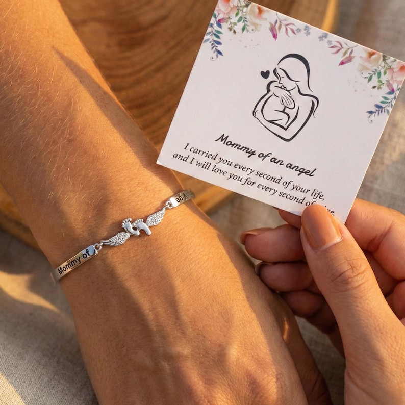 May include: A silver bracelet with the words "Mommy of" engraved on it. The bracelet features a charm with baby footprints and angel wings. A card with the text "Mommy of an angel" and a mother and child illustration is held nearby.