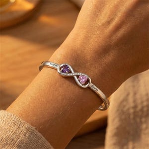May include: A silver-toned cuff bracelet featuring an infinity symbol design. The bracelet has two heart-shaped gemstones, one purple and one pink, connected by a row of small, clear stones. The names "Kaitlyn" and "Evelyn" are engraved on the bracelet.