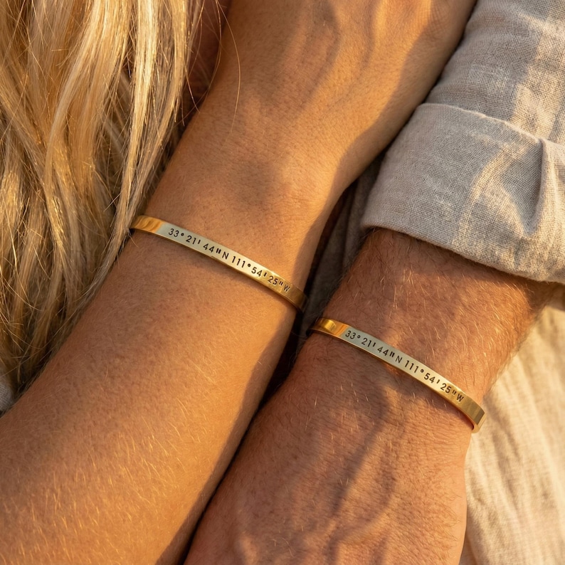 May include: Two gold-colored metal bracelets, each engraved with the coordinates "33° 21' 44" N 111° 54' 25" W". The bracelets are simple bands, worn on wrists. The image shows a close-up of the bracelets.