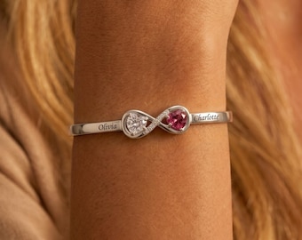 May include: A silver-toned bangle bracelet featuring an infinity symbol centerpiece. The symbol is accented with a clear heart-shaped stone and a pink heart-shaped stone. The names Olivia and Charlotte are engraved on the metal band.