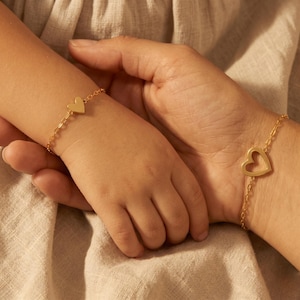 May include: Two gold-colored heart bracelets. One bracelet is on a child's wrist, featuring a solid heart charm. The other bracelet, on an adult's wrist, has a heart-shaped cutout. Both bracelets are on delicate chain links.