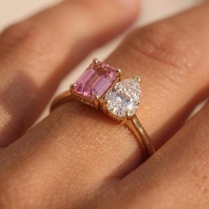 Op de afbeelding: Een gouden ring met twee stenen naast elkaar: een rechthoekige roze edelsteen met smaragdgeslepen facetten en een peervormige helderwitte edelsteen. De ringband is van gepolijst goud en de stenen worden vastgehouden door kleine gouden pootjes.