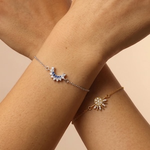Puede incluir: Una pulsera de cadena plateada con un dije de media luna engastado con piedras azules y transparentes, y una pulsera de cadena dorada con un dije en forma de sol engastado con piedras transparentes, ambas lucidas en las muñecas cruzadas.