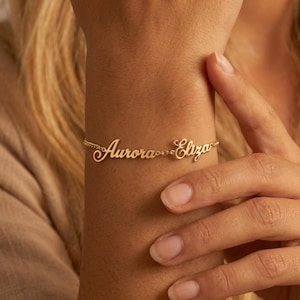 Op de afbeelding: Een goudkleurige metalen armband met de twee namen Aurora en Eliza in een cursief lettertype. De namen zijn verbonden door een dunne, delicate gouden ketting die om de pols wikkelt, wat zorgt voor een gepersonaliseerd sieraad.