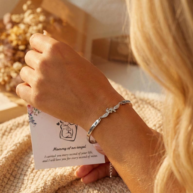 May include: A silver bracelet with the words "Mommy of" engraved on it. The bracelet features a charm with angel wings and small crystals. A card with the text "Mommy of an angel" is held in the background.
