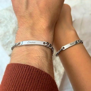 Könnte beinhalten: Zwei silberfarbene Kettenarmbänder. Eines hat den Namen "Emma" auf einer rechteckigen Platte eingraviert. Das andere hat den Namen "Mert". Die Armbänder werden an Handgelenken getragen.