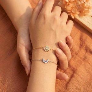 May include: Two delicate bracelets adorn a wrist. The top bracelet is gold with a sunburst design, while the bottom is silver with a crescent moon shape. Both bracelets are embellished with small, sparkling stones.