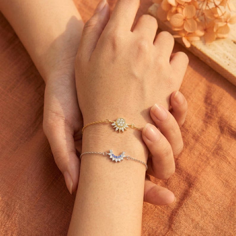 May include: Two delicate bracelets adorn a wrist. The top bracelet is gold with a sunburst design, while the bottom is silver with a crescent moon shape. Both bracelets are embellished with small, sparkling stones.
