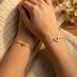 May include: Two gold-colored heart bracelets. One bracelet features a solid heart charm, and the other has a heart-shaped cutout. The bracelets are on delicate chains. The background is a neutral beige.