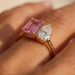 Pode incluir: Um anel de ouro com duas pedras proeminentes: uma gema rosa de corte esmeralda retangular e uma gema branca transparente em forma de pera. As pedras estão engastadas lado a lado num aro de ouro liso e polido que envolve o dedo.