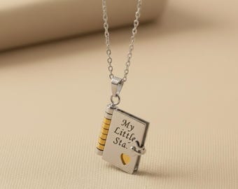 May include: Silver-tone necklace with a pendant shaped like a small book. The book pendant has the words "My Little Sta." engraved on the cover, with a heart-shaped cutout. The spine is gold and the pages are silver.