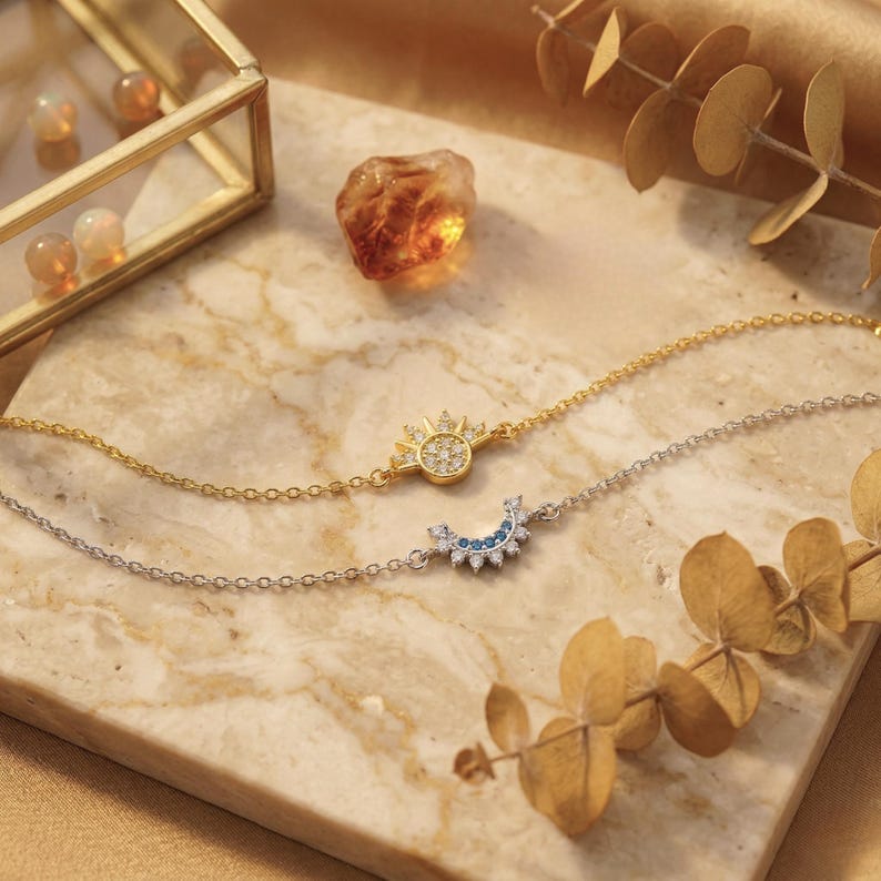 May include: Two delicate chain bracelets. One is gold-toned with a sunburst pendant, the other is silver-toned with a crescent moon pendant. Both are adorned with small, sparkling stones. The bracelets rest on a marble surface.