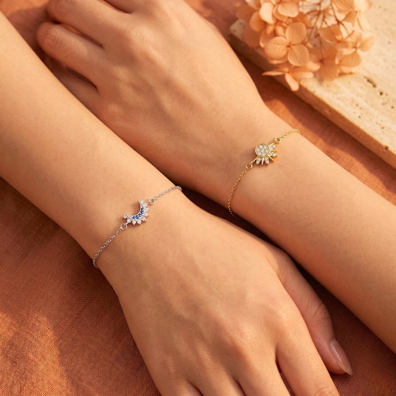 May include: Two bracelets, one silver and one gold, are displayed on wrists. The silver bracelet features a crescent moon design with blue and clear stones. The gold bracelet has a sun-shaped charm with clear stones. The background is a warm, neutral tone.