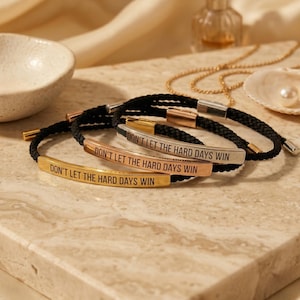 Könnte beinhalten: Drei Armbänder mit der Aufschrift "DON'T LET THE HARD DAYS WIN". Die Armbänder haben ein schwarzes Seildesign mit goldenen, silbernen und roségoldenen Metallakzenten. Sie werden auf einer Marmoroberfläche präsentiert.