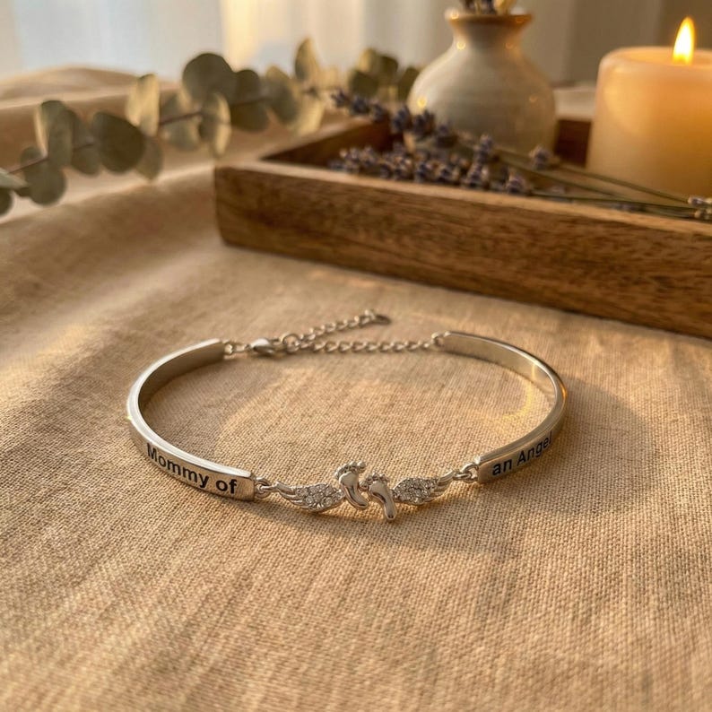 May include: Silver-toned bracelet with the words "Mommy of an Angel" engraved on it. The bracelet features a decorative centerpiece with two angel figures and small, clear stones. The bracelet is displayed on a beige surface.