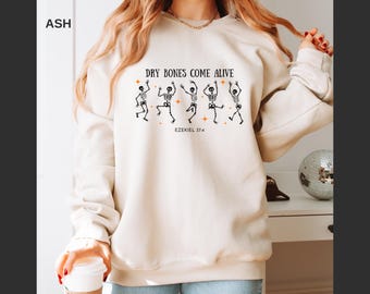 May include: Ash-colored sweatshirt featuring a graphic of dancing skeletons with the text "DRY BONES COME ALIVE" and "EZEKIEL 37:4". The design includes orange star accents. The sweatshirt is a neutral color.