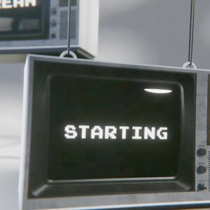 May include: A vintage-style television with a black and white screen displaying the word "STARTING" in white pixelated letters. The television is hanging from the ceiling and has a wooden frame with silver knobs.