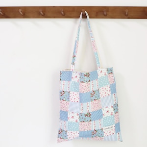 May include: A patchwork tote bag with a floral and gingham pattern in shades of blue and pink. The bag hangs from a wooden rack with pegs. The bag has long straps and a rectangular shape.