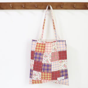 May include: A patchwork tote bag with a variety of floral and plaid patterns in shades of red, pink, yellow, blue, and white. The bag has a white canvas strap and a small label that reads "LAVENDER".